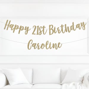 Peut inclure: Une banni&egrave;re dor&eacute;e &agrave; paillettes avec l'inscription "Happy 21st Birthday Caroline" est suspendue sur un mur blanc. La banni&egrave;re est au-dessus d'un canap&eacute; blanc avec des coussins blancs. Le texte est en &eacute;criture cursive.