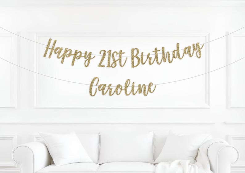 Peut inclure: Une banni&egrave;re dor&eacute;e &agrave; paillettes avec l'inscription "Happy 21st Birthday Caroline" est suspendue sur un mur blanc. La banni&egrave;re est au-dessus d'un canap&eacute; blanc avec des coussins blancs. Le texte est en &eacute;criture cursive.