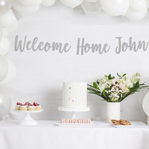 May include: A white table with a white tablecloth, decorated with a silver glitter banner that says "Welcome Home John". There are white balloons, a white cake with gold sprinkles, cupcakes with strawberries, pink macarons, and cookies on the table.