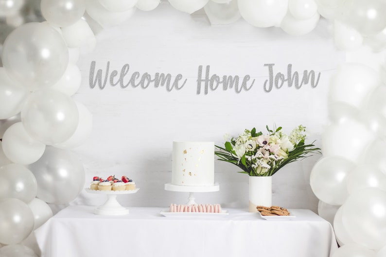 May include: A white table with a white tablecloth, decorated with a silver glitter banner that says "Welcome Home John". There are white balloons, a white cake with gold sprinkles, cupcakes with strawberries, pink macarons, and cookies on the table.