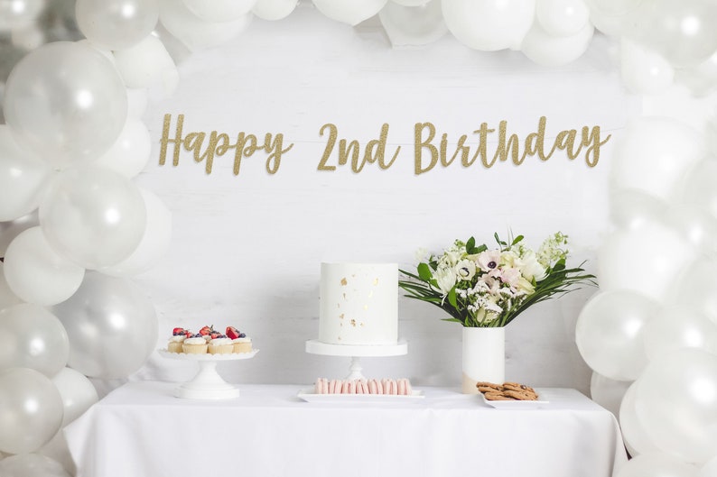 Op de afbeelding: Een witte tafel met een wit tafelkleed, een witte taart met gouden sprinkles, cupcakes met bessen, een boeket witte bloemen en een gouden glitterbanner met de tekst "Happy 2nd Birthday".