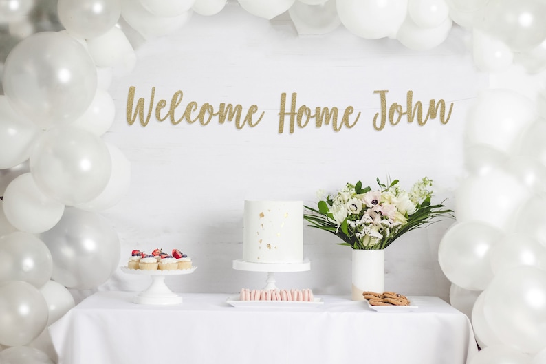 May include: A white table with a white tablecloth, a white cake with gold flecks, cupcakes with red berries, a bouquet of white flowers in a white vase, and a gold glitter banner that reads "Welcome Home John".