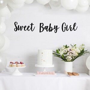 May include: White table with a white tablecloth, a white cake with gold flecks, cupcakes with red berries, pink macarons, and a bouquet of white flowers in a white vase. A white balloon arch is in the background with a black banner that reads "Sweet Baby Girl".