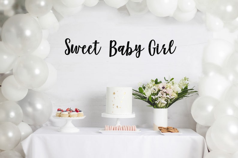 May include: White table with a white tablecloth, a white cake with gold flecks, cupcakes with red berries, pink macarons, and a bouquet of white flowers in a white vase. A white balloon arch is in the background with a black banner that reads "Sweet Baby Girl".