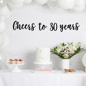 May include: A white table with a white tablecloth, a white cake with gold sprinkles, cupcakes with red berries, pink macarons, and cookies. A bouquet of white and pink flowers in a white vase is on the table. A white balloon arch is in the background. The text "Cheers to 30 years" is written in black on the wall.