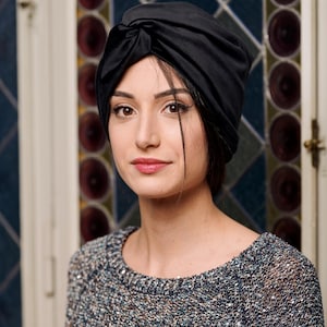 Può includere: Una donna indossa un turbante nero e un maglione grigio scintillante. È in piedi davanti a una finestra con vetri colorati.