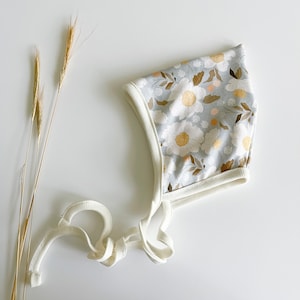 May include: A white baby bonnet with a floral pattern in shades of brown, yellow, and white on a light blue background. The bonnet has a white trim and ties.