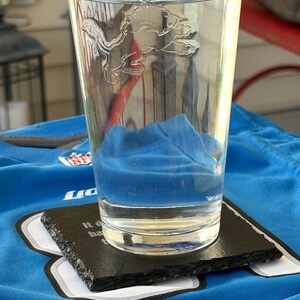 May include: A clear glass with a Detroit Lions logo etched on the side, filled with water, sits on a black slate coaster. The coaster rests on a blue jersey. A blue object is submerged in the water.
