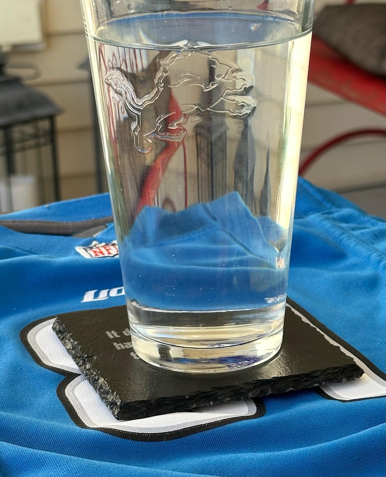 May include: A clear glass with a Detroit Lions logo etched on the side, filled with water, sits on a black slate coaster. The coaster rests on a blue jersey. A blue object is submerged in the water.