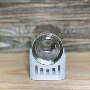 May include: A clear glass shot glass sits upside down on a white plastic holder with a grid pattern.