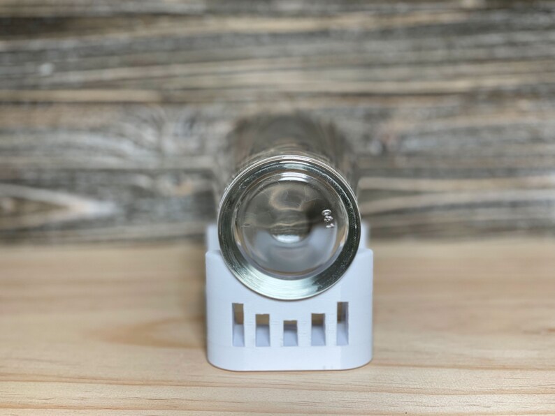 May include: A clear glass shot glass sits upside down on a white plastic holder with a grid pattern.