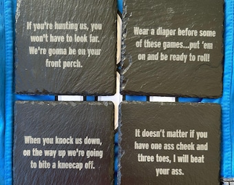 May include: Four square black slate coasters with white engraved text. The coasters have various phrases, including "If you're hunting us..." and "Wear a diaper before some of these games..." The coasters are on a blue surface with the word "LIONS" visible.