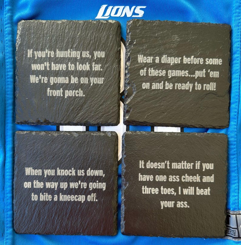 May include: Four square black slate coasters with white engraved text. The coasters have various phrases, including "If you're hunting us..." and "Wear a diaper before some of these games..." The coasters are on a blue surface with the word "LIONS" visible.