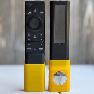 May include: A black Samsung television remote control and a black solar-powered remote control, both inserted into bright yellow plastic stands. The right stand features a circular silver Apple AirTag with a visible logo on its front surface.