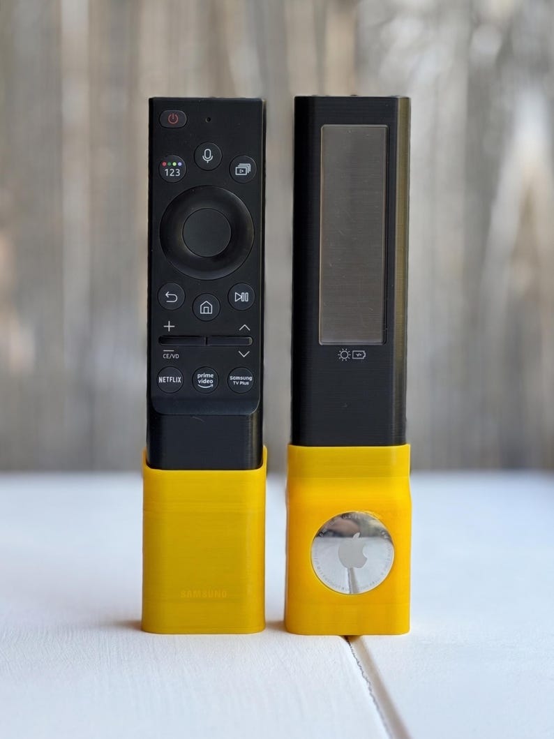 Puede incluir: Un mando a distancia de televisi&oacute;n Samsung negro y un mando a distancia solar negro, ambos insertados en soportes de pl&aacute;stico amarillo brillante. El soporte derecho cuenta con un Apple AirTag plateado circular con un logotipo visible en su parte frontal.