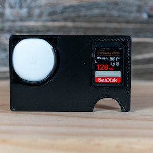 May include: A black plastic case with a white round object and a SanDisk Extreme PRO 128 GB SD card.