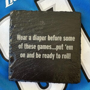May include: A square, black slate coaster with the engraved text: "Wear a diaper before some of these games...put 'em on and be ready to roll!" The coaster has a rough, textured surface and is likely used for beverages.