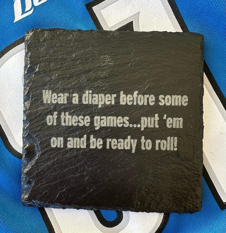 May include: A square, black slate coaster with the engraved text: "Wear a diaper before some of these games...put 'em on and be ready to roll!" The coaster has a rough, textured surface and is likely used for beverages.