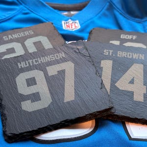 May include: Four slate coasters with player names and numbers, including "SANDERS 20", "HUTCHINSON 97", "ST. BROWN 14", and "GOFF". The coasters are dark gray and rest on a blue jersey with an NFL logo.