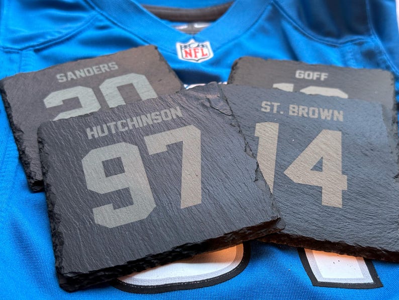 May include: Four slate coasters with player names and numbers, including "SANDERS 20", "HUTCHINSON 97", "ST. BROWN 14", and "GOFF". The coasters are dark gray and rest on a blue jersey with an NFL logo.