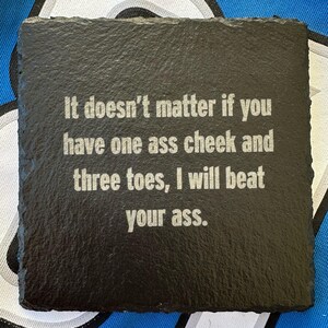 May include: Square black slate coaster with the text "It doesn't matter if you have one ass cheek and three toes, I will beat your ass." in a white, sans-serif font. The coaster has a rough, textured surface.