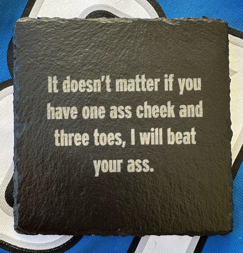 May include: Square black slate coaster with the text "It doesn't matter if you have one ass cheek and three toes, I will beat your ass." in a white, sans-serif font. The coaster has a rough, textured surface.