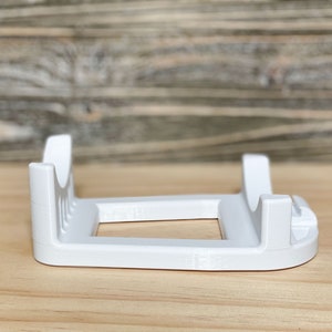 May include: White plastic stand with two slots for holding items. The stand has a rectangular base and two vertical supports.