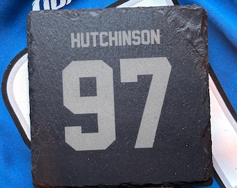 May include: Square black slate coaster with the name "HUTCHINSON" and the number "97" in gray lettering. The coaster is resting on a blue jersey with the word "LIONS" visible.