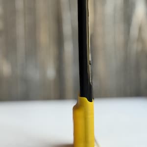 Puede incluir: Una herramienta negra, delgada y rectangular con un acabado liso, insertada en una base de pl&aacute;stico moldeado de color amarillo brillante. La base presenta una forma contorneada que sujeta la parte inferior del objeto negro en posici&oacute;n vertical.