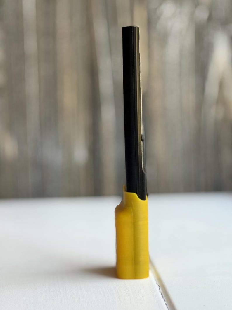 Puede incluir: Una herramienta negra, delgada y rectangular con un acabado liso, insertada en una base de pl&aacute;stico moldeado de color amarillo brillante. La base presenta una forma contorneada que sujeta la parte inferior del objeto negro en posici&oacute;n vertical.