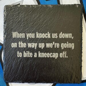 May include: A square, black slate coaster with a rough texture. The coaster features the engraved text: "When you knock us down, on the way up we're going to bite a kneecap off."