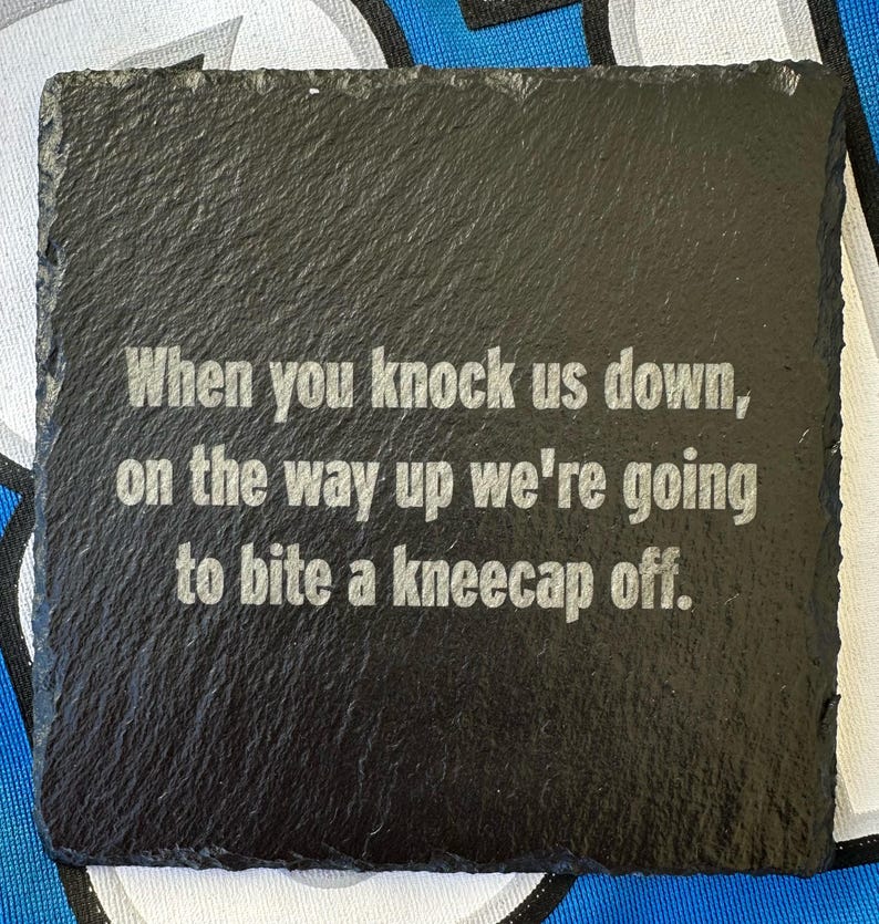 May include: A square, black slate coaster with a rough texture. The coaster features the engraved text: "When you knock us down, on the way up we're going to bite a kneecap off."