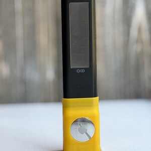 Puede incluir: Un dispositivo electr&oacute;nico rectangular negro con un panel solar y un icono de bater&iacute;a impreso en el frente. La parte inferior est&aacute; encajada en un soporte de pl&aacute;stico amarillo brillante con un recorte circular que muestra un Apple AirTag plateado.