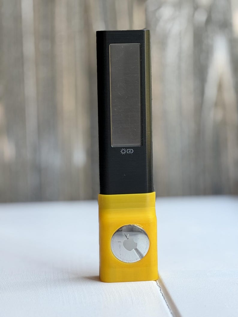 Puede incluir: Un dispositivo electr&oacute;nico rectangular negro con un panel solar y un icono de bater&iacute;a impreso en el frente. La parte inferior est&aacute; encajada en un soporte de pl&aacute;stico amarillo brillante con un recorte circular que muestra un Apple AirTag plateado.