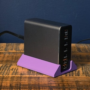Könnte beinhalten: Ein schwarzes Multi-Port-USB-Ladegerät mit lila Basis, das auf einer Holzoberfläche steht. Das Ladegerät verfügt über mehrere USB-C- und USB-A-Anschlüsse. Ein schwarzes Netzkabel ist eingesteckt.