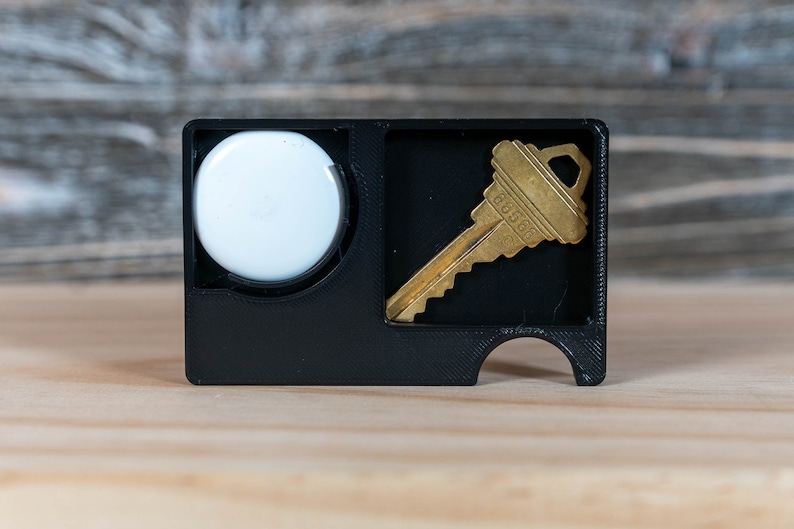 May include: A black plastic key holder with a white circle and a gold key inside. The key holder has a slot for a key and a small hole for a keychain.