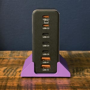 May include: A black USB charging station with multiple ports, including USB-C and USB-A, is displayed in a purple stand. The ports are labeled USB-C1 through USB-C5 and USB-A1 and USB-A2. The charger is on a wooden surface.