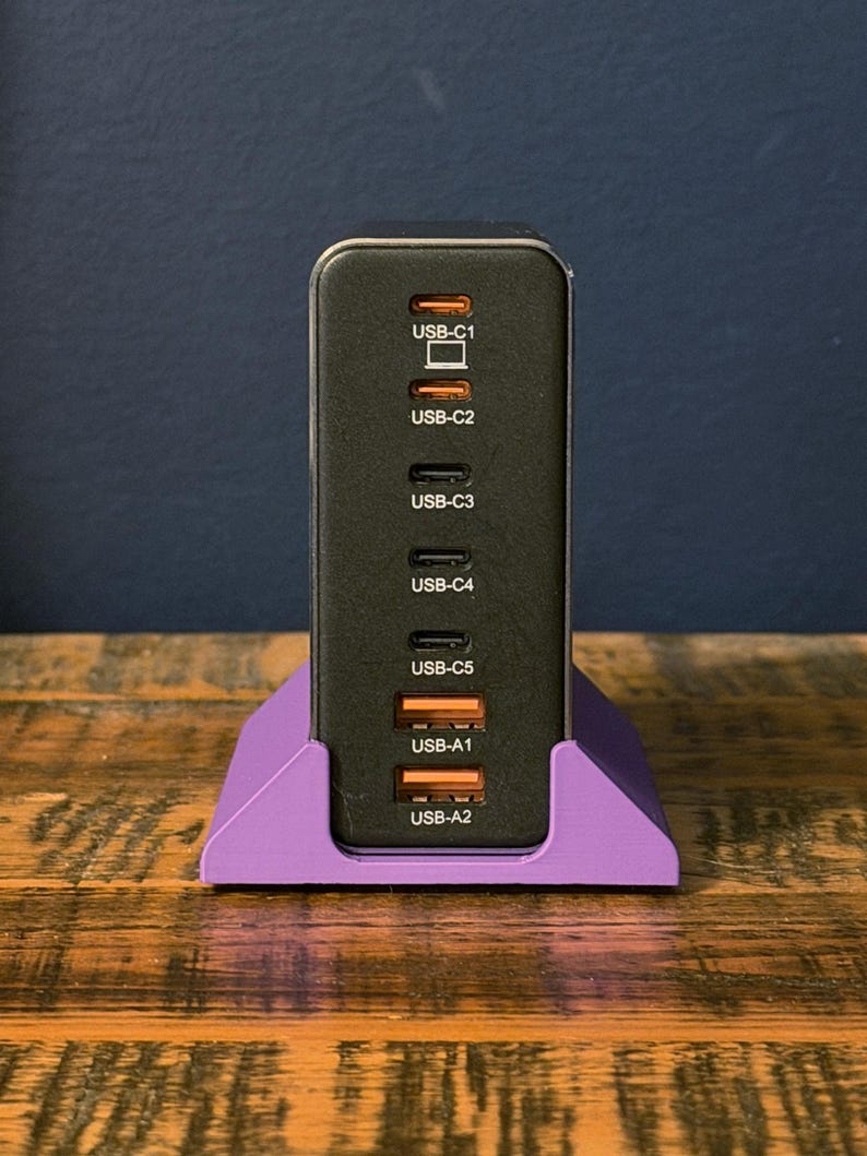 May include: A black USB charging station with multiple ports, including USB-C and USB-A, is displayed in a purple stand. The ports are labeled USB-C1 through USB-C5 and USB-A1 and USB-A2. The charger is on a wooden surface.