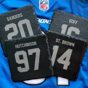 May include: Four square black slate coasters with player names and numbers: Sanders 20, Goff 16, Hutchinson 97, and St. Brown 14. The coasters are displayed on a blue jersey with an NFL logo.