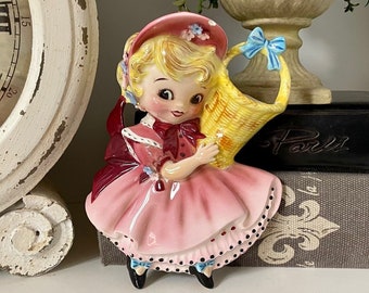 Puede incluir: Una figurita de pared vintage de cerámica de una niña con un vestido rosa y un sombrero azul, sosteniendo una cesta amarilla. La figurita se exhibe en un estante con un libro titulado "París 1898" y un libro negro con la palabra "París" en él.