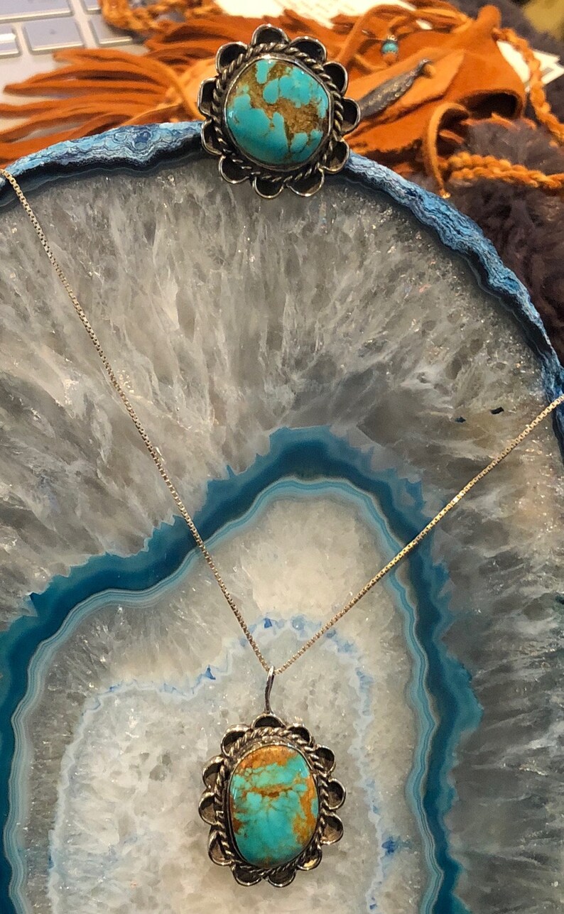 May include: A turquoise and silver pendant necklace and ring set. The pendant and ring feature a turquoise stone with brown veins and a silver setting with a scalloped edge.