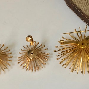 May include: A set of three gold-toned jewelry pieces featuring a sunburst design. The set includes a pair of clip-on earrings and a brooch.