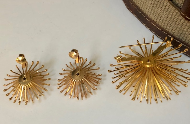 May include: A set of three gold-toned jewelry pieces featuring a sunburst design. The set includes a pair of clip-on earrings and a brooch.