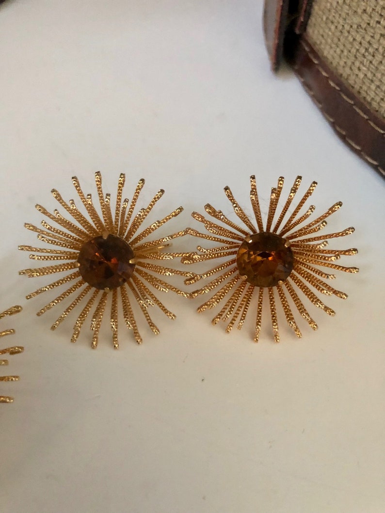May include: A pair of gold tone earrings with a starburst design and amber colored rhinestones in the center.