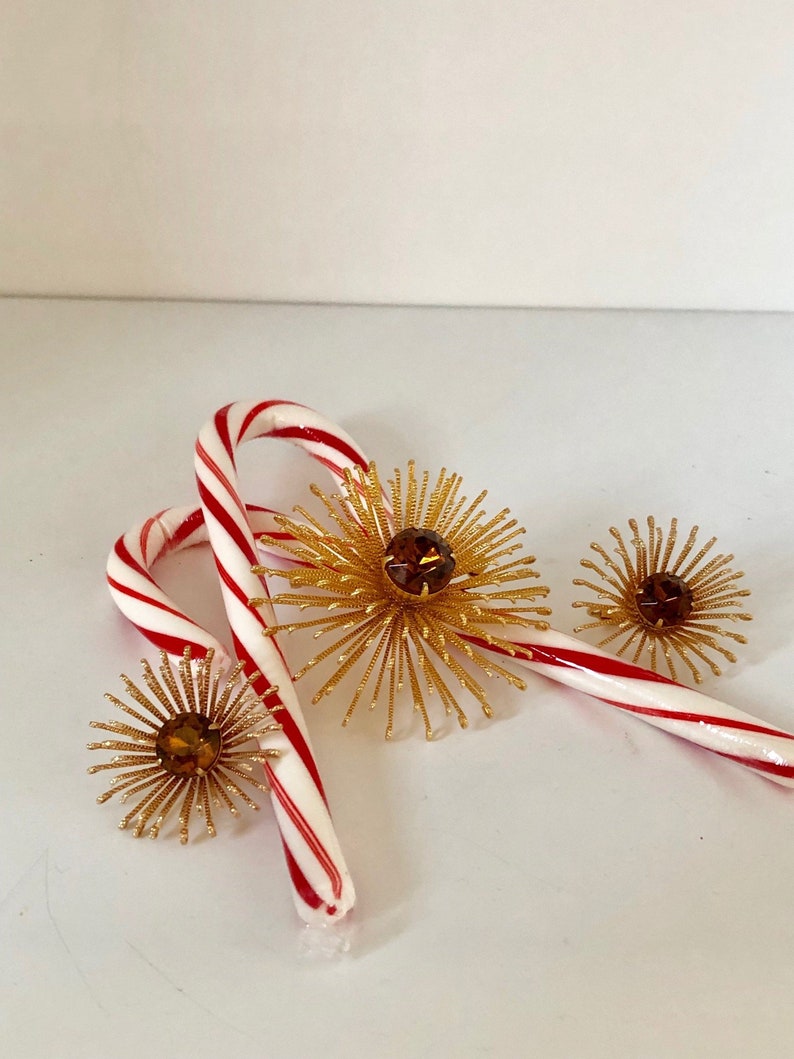 May include: Three gold starburst ornaments with amber colored rhinestones in the center. The ornaments are sitting on top of two red and white candy canes.