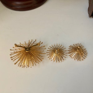 May include: A set of three gold-toned jewelry pieces. The largest piece is a brooch with a starburst design. The two smaller pieces are earrings with a similar starburst design.