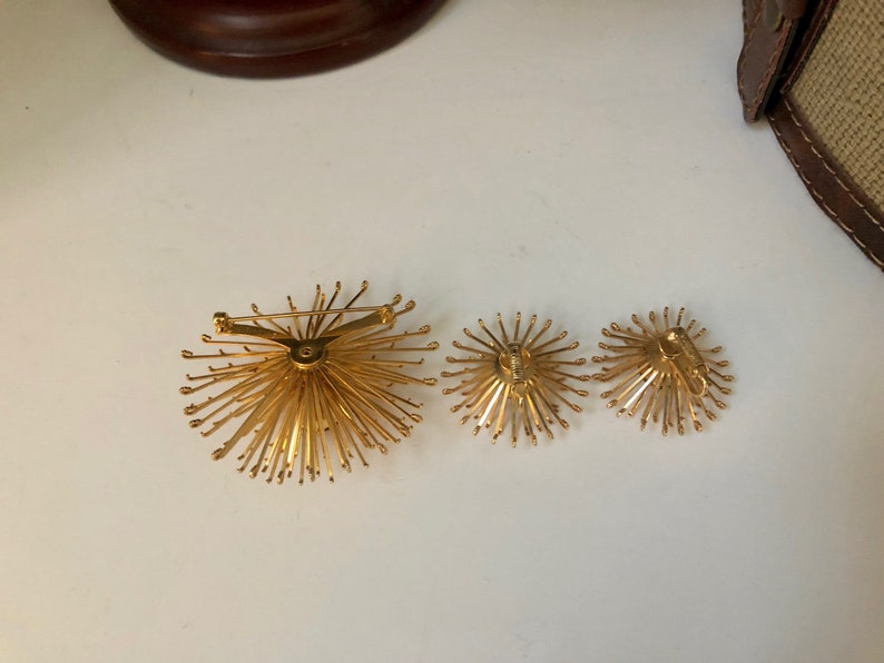 May include: A set of three gold-toned jewelry pieces. The largest piece is a brooch with a starburst design. The two smaller pieces are earrings with a similar starburst design.