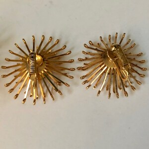 May include: A pair of gold tone clip-on earrings with a sunburst design. The earrings are made of metal and have a circular shape. The earrings are about 1 inch in diameter.