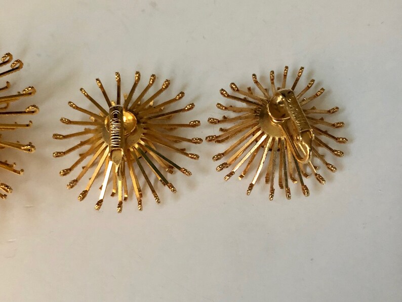 May include: A pair of gold tone clip-on earrings with a sunburst design. The earrings are made of metal and have a circular shape. The earrings are about 1 inch in diameter.
