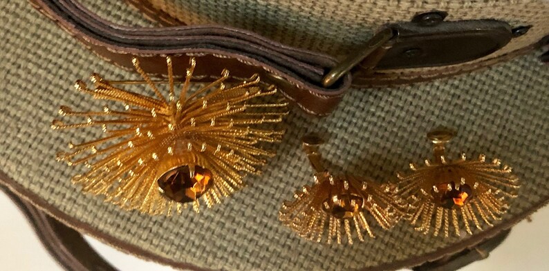 May include: A pair of gold tone earrings with a large, round, amber-colored stone in the center of each earring. The earrings are surrounded by a burst of gold-colored metal.